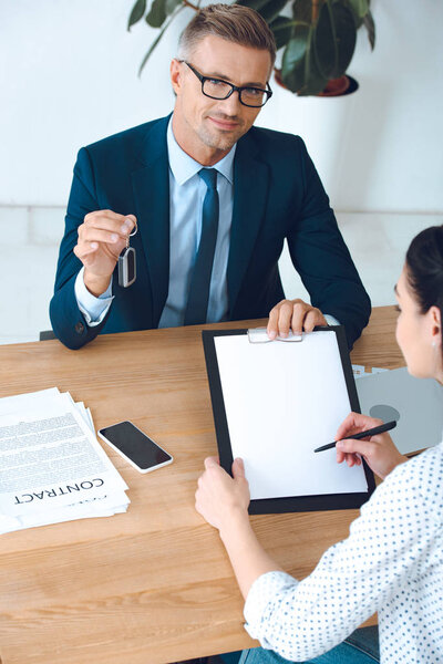 high angle view of smiling insurance agent with car key and client signing papers at workplace in office