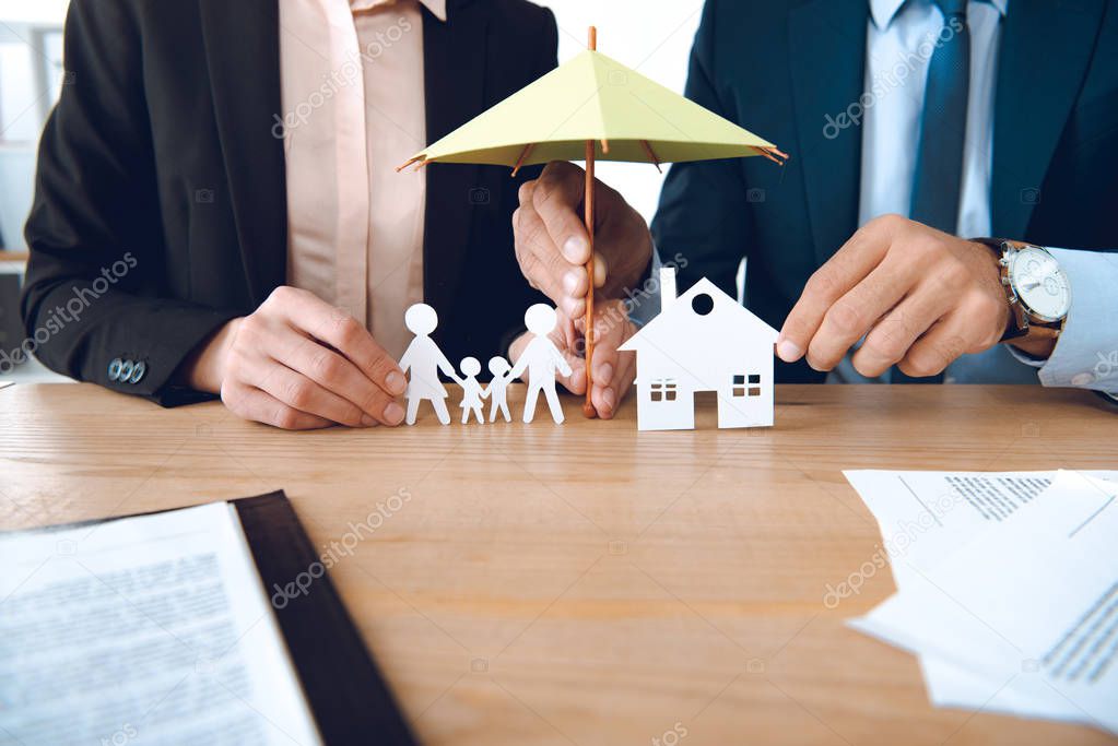 partial view of insurance agents covering family and house paper models with umbrella at workplace