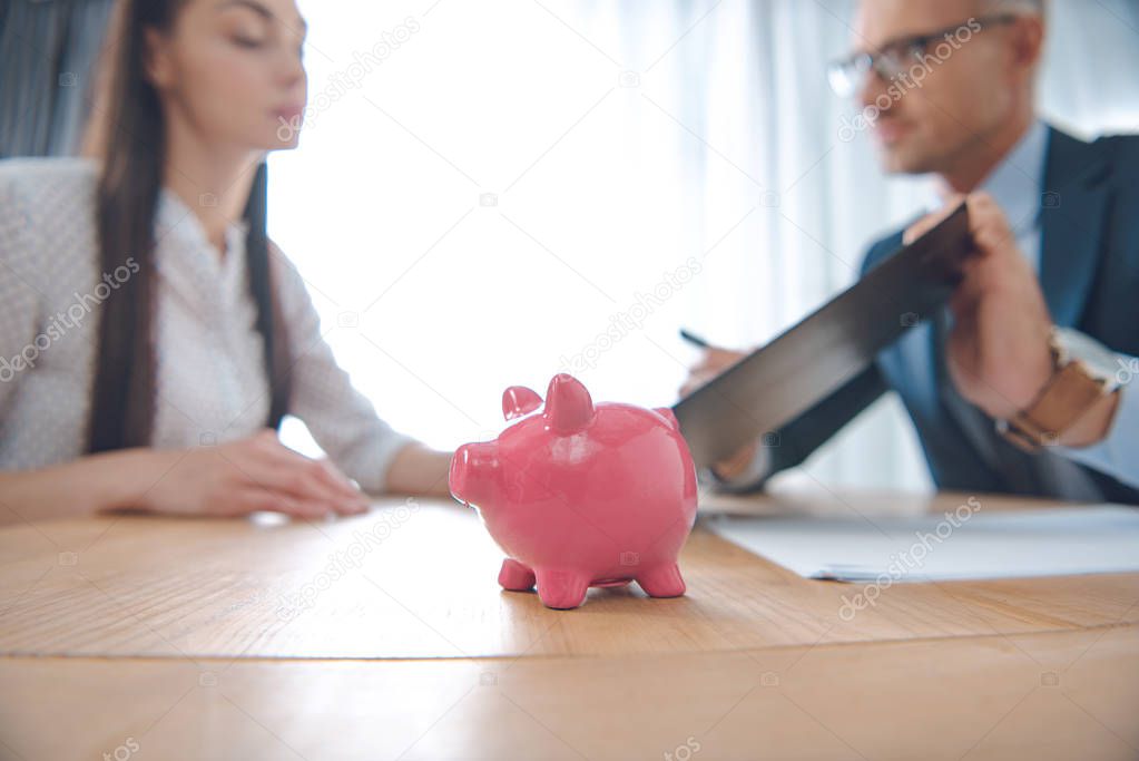selective focus of insurance agent and client at tabletop with pink piggy bank