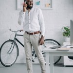 Усміхнений красивий афроамериканський бізнесмен розмовляє по смартфону в офісі і дивиться геть