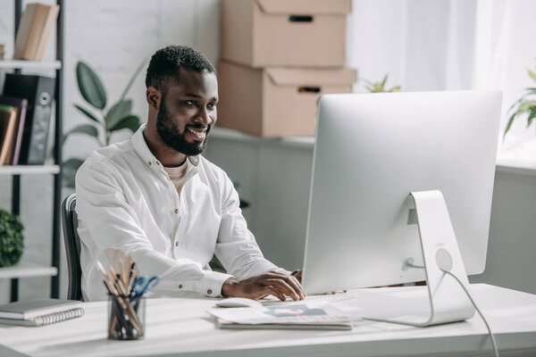 smiling handsome african american businessman in white shirt working at computer in office