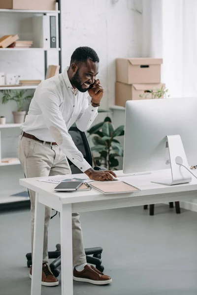 笑顔のハンサムなアフリカ系アメリカ人のビジネスマンはスマート フォンで話していると オフィスのコンピューターを使用して  — 無料ストックフォト