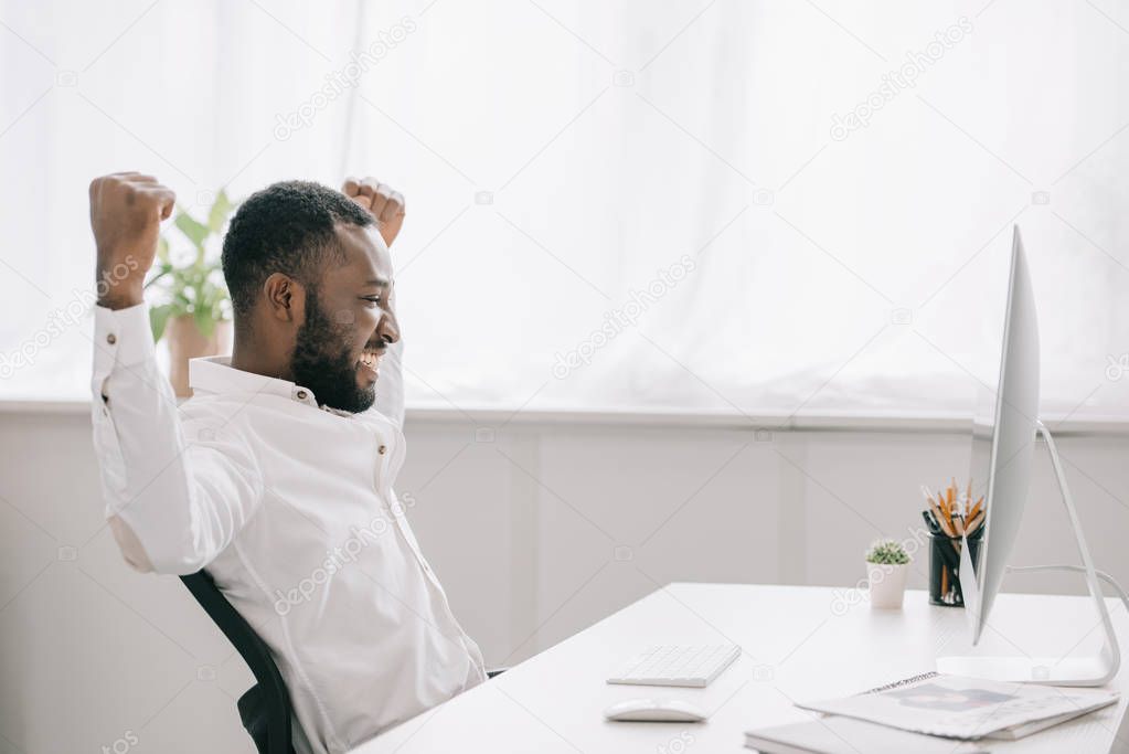 side view of happy african american businessman working at computer in office and showing yes gesture