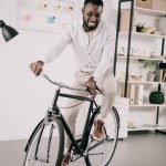 Sonriente guapo afroamericano hombre de negocios montar en bicicleta en la oficina