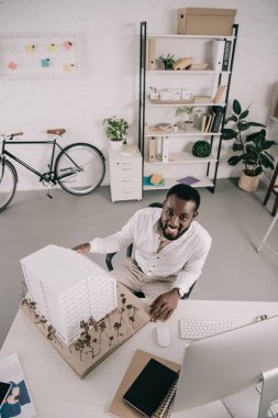 gülümseyen yakışıklı Afrikalı-Amerikalı mimar mimari modeli oturan ve kamera Office bakarak yüksek açılı görünüş