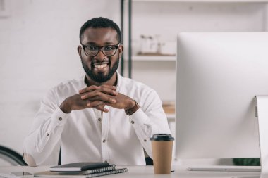 masada oturan ve kamera Office bakarak gülümseyen yakışıklı Afrikalı-Amerikalı işadamı