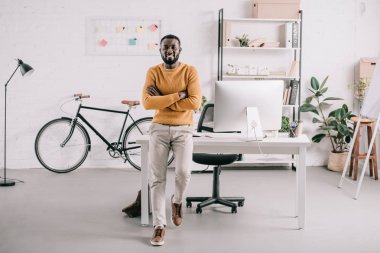 masanın üzerine eğilerek ve kamera Office bakarak çapraz kollu yakışıklı Afro-Amerikan Tasarımcısı