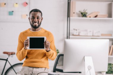gülümseyen yakışıklı Afrikalı-Amerikalı tasarımcı turuncu kazaklı ofiste tablet boş ekran ile gösterilen