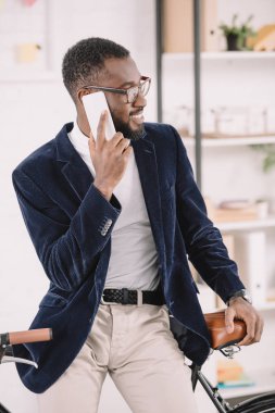 neşeli Afro-Amerikan Yöneticisi smartphone üzerinde bisiklet Office yaslanmış iken konuşuyor