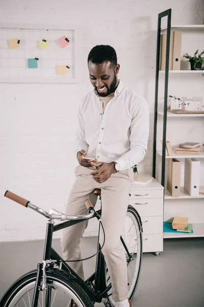 Sonriente Guapo Afroamericano Hombre Negocios Sentado Bicicleta Uso Smartphone Oficina — Foto de stock gratis