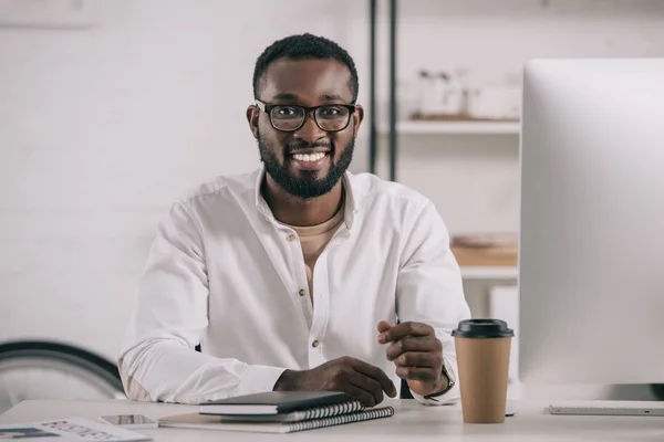 ガラスのテーブルに座って オフィスでのカメラ目線の笑顔のハンサムなアフリカ系アメリカ人実業家 — ストック写真