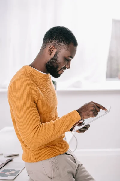 英俊的非洲美国设计师在橙色毛衣的侧面视图使用平板电脑在办公室 — 免费的图库照片