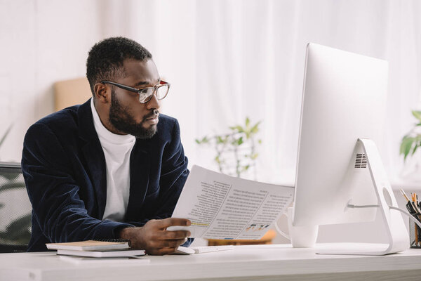 bearded african american businessman holding document and working with computer at workplace