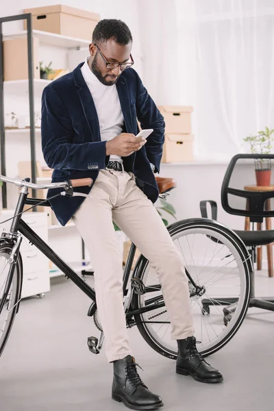アフリカ系アメリカ人のビジネスマンがオフィスで自転車にもたれながらスマート フォンを使用してひげを生やした — ストック写真