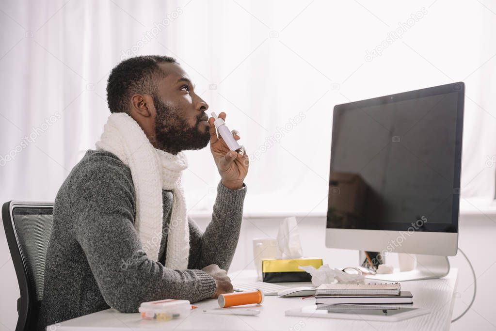 ill african american businessman in warm scarf with nose spray at workplace with computer