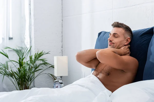 shirtless adult man with closed eyes holding hands behind neck in bed at home