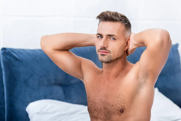 portrait of man with hands behind neck while sitting on bed at home