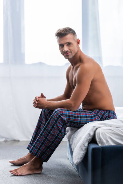 smiling shirtless adult man sitting on bed during morning time at home