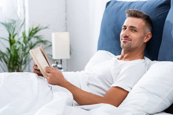 happy man reading book while laying in bed during morning time at home