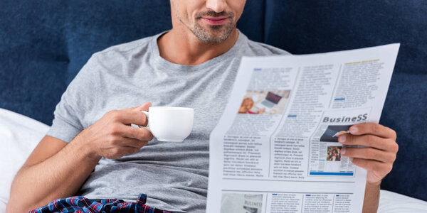 partial view of businessman with cup of coffee reading business newspaper in bed at home