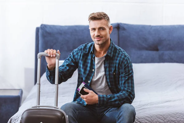 Happy Male Traveler Sitting Bed Smartphone Flight Ticket Wheeled Bag — Free Stock Photo