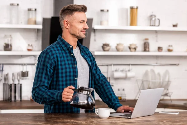 Knappe Man Bedrijf Koffie Pot Lachend Met Behulp Van Laptop — Gratis stockfoto