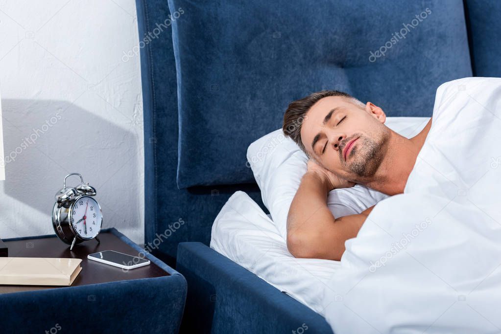 adult man sleeping in bed near alarm clock and smartphone with blank screen at home