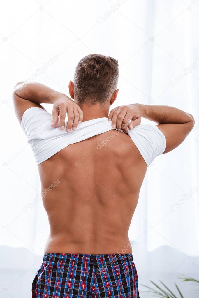 back view of man putting on white t-shirt while sitting on bed at home