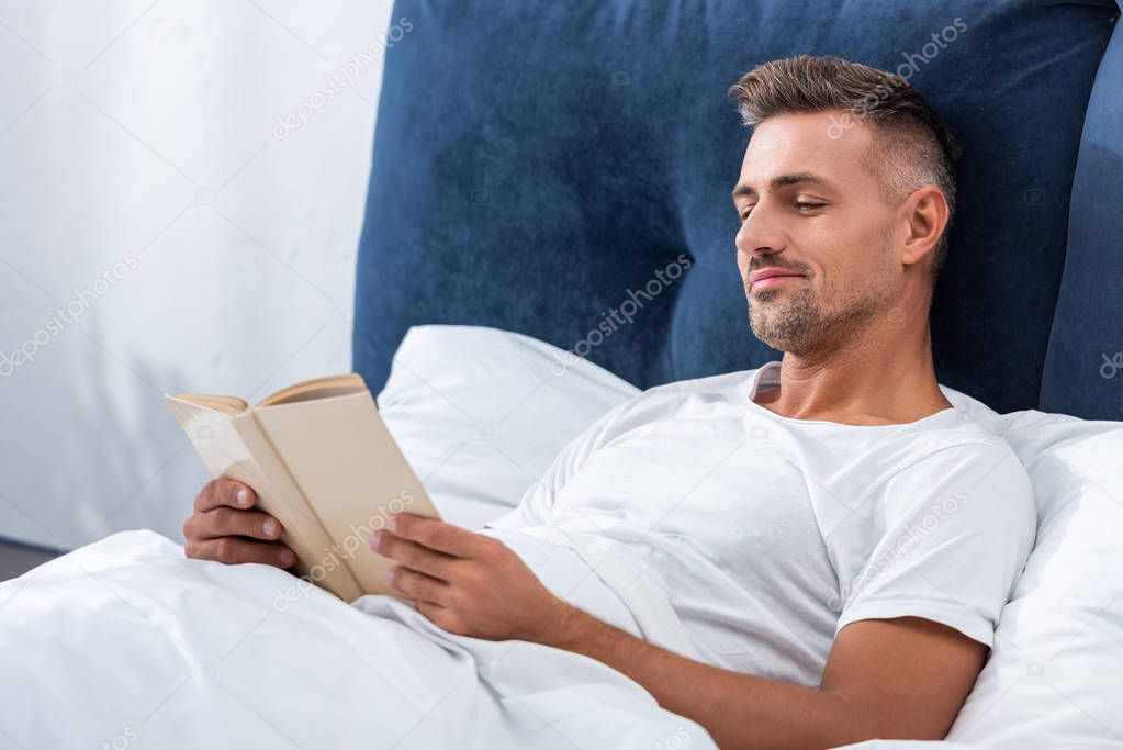 smiling man reading book while laying in bed during morning time at home