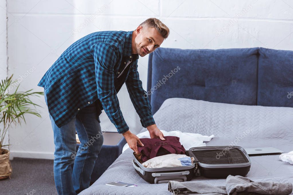 smiling male traveler packing luggage in bedroom at home 