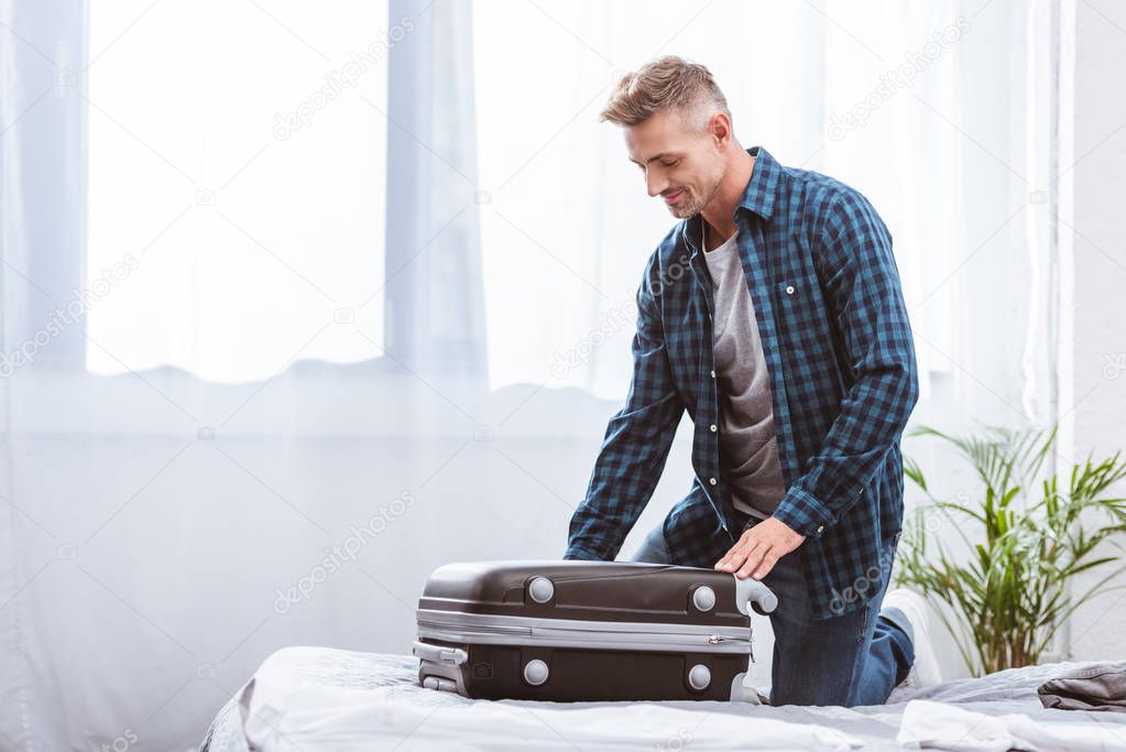 cheerful male traveler packing travel bag in bedroom at home