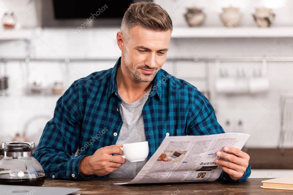 smiling man in checkered shirt holding cup of coffee and reading newspaper at home