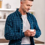 Bel homme souriant en chemise à carreaux tenant une tasse de café à la maison