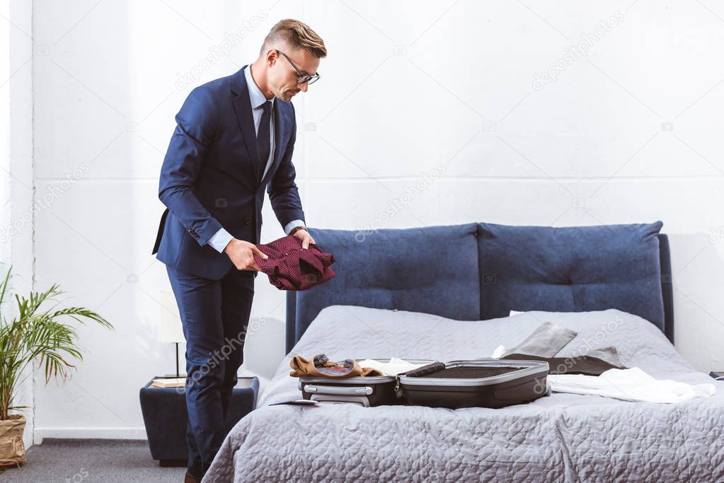 handsome businessman in suit and eyeglasses packing suitcase in bedroom