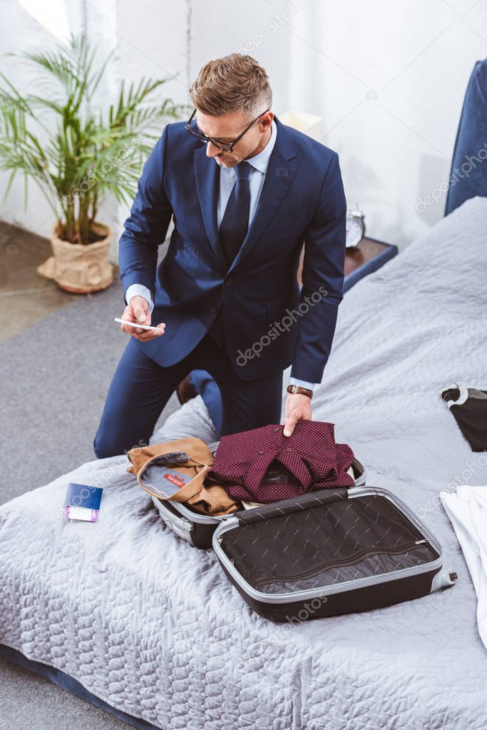 high angle view of man in suit and eyeglasses using smartphone and packing suitcase for business trip