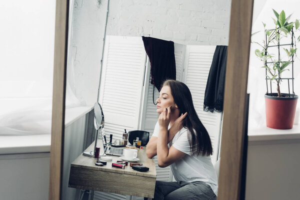 view through mirror reflection at young transgender woman doing makeup at home