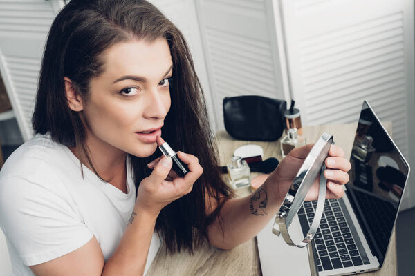 close-up shot of young transgender woman applying lipstick while looking at camera