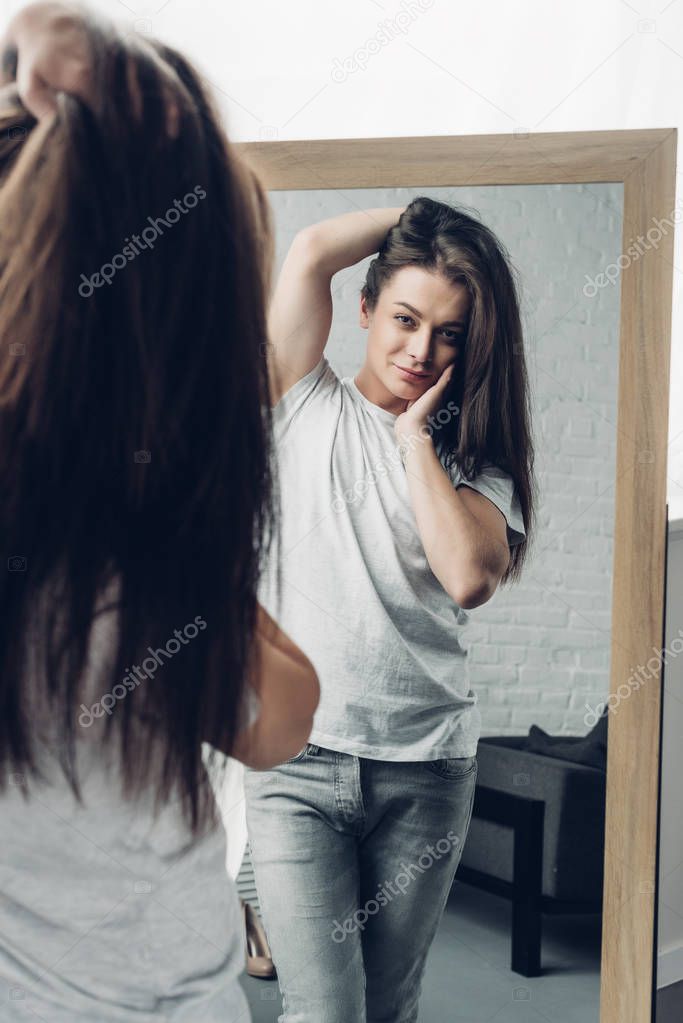 young transgender woman in white t-shirt looking at mirror at home