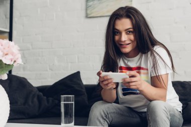 smiling young transgender woman using smartphone on couch at home clipart