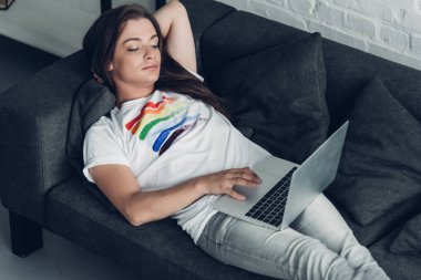 relaxed young transgender freelancer woman working with laptop on couch at home clipart