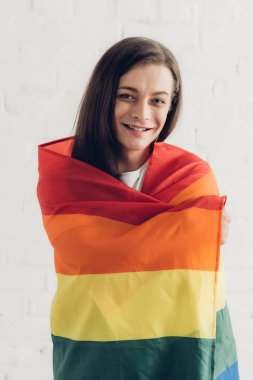 happy transgender woman covering with pride flag and looking at camera in front of white brick wall clipart