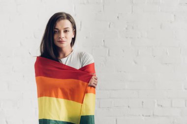 Genç transseksüel kadın gurur bayrağıyla örtünüyor ve beyaz tuğla duvarın önünde kameraya bakıyor.