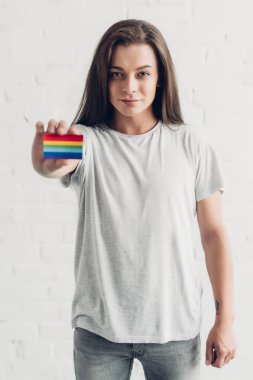 young transgender woman holding card with pride flag in front of white brick wall clipart