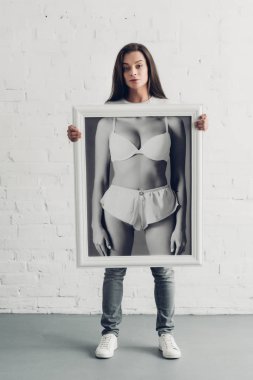 young transgender woman holding black and white photo with female body in underwear in front of white brick wall clipart