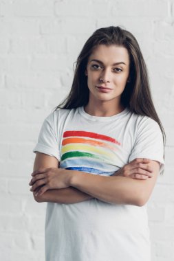 young transgender woman in white t-shirt with pride flag looking at camera with crossed arms in front of white brick wall clipart