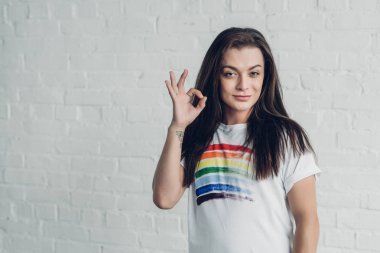 young transgender woman in white t-shirt with pride flag showing okay sign in front of white brick wall clipart