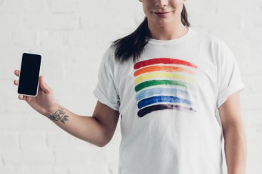 cropped shot of young transgender woman in white t-shirt with pride flag holding smartphone with blank screen in front of white brick wall clipart