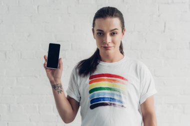 Beyaz tişörtlü genç transseksüel kadın gururlu bayraklı beyaz duvarın önünde beyaz ekranlı akıllı telefon tutuyor.