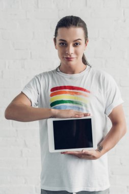 Beyaz tişörtlü genç transseksüel kadın beyaz tuğla duvarın önünde boş ekranlı tablet taşıyor.