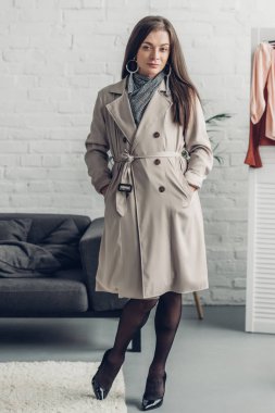 young transgender woman in trench coat looking at camera at home clipart